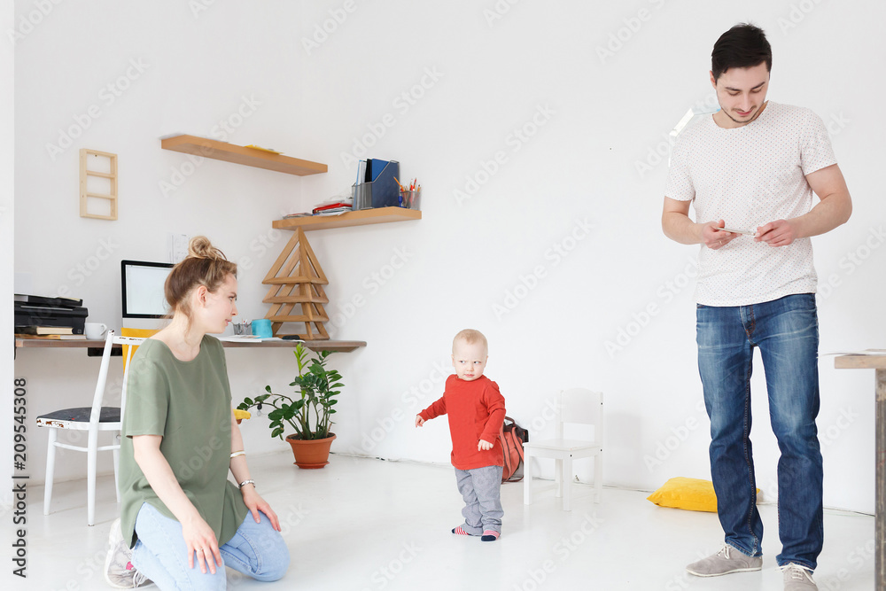 First steps. Young mother cute dad watching how their little child takes the first steps in the room in modern interior. Copyspace First steps. Young mother cute dad watching how their little child takes the first steps in the room in modern interior. Copyspace