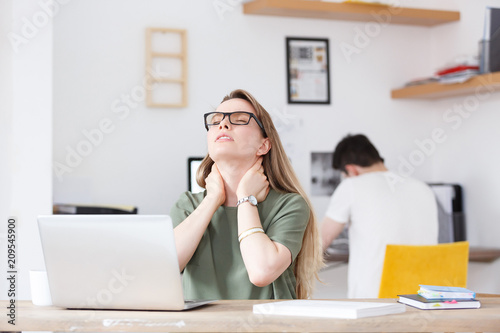 Pain in the neck. Beautiful young woman rubbed tired at the office sitting at table on background of blurred man employee