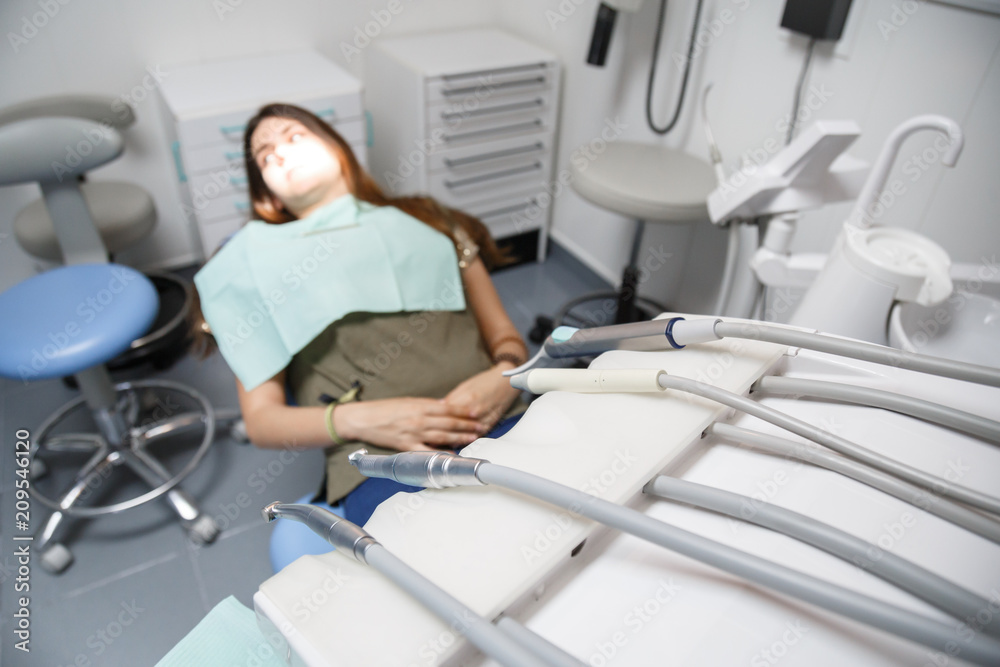 Young female sitting in dental chair in clinic room with equipment in soft focus. 