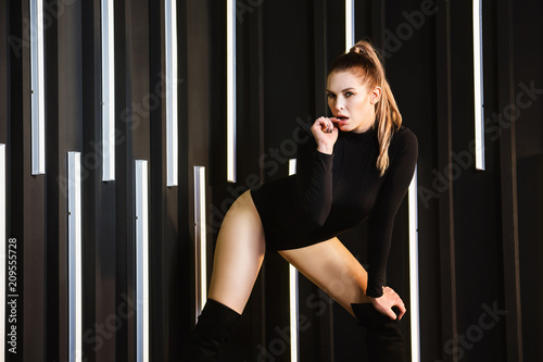 Sexy blond girl in body shirt and boots is posing at dark wall background.