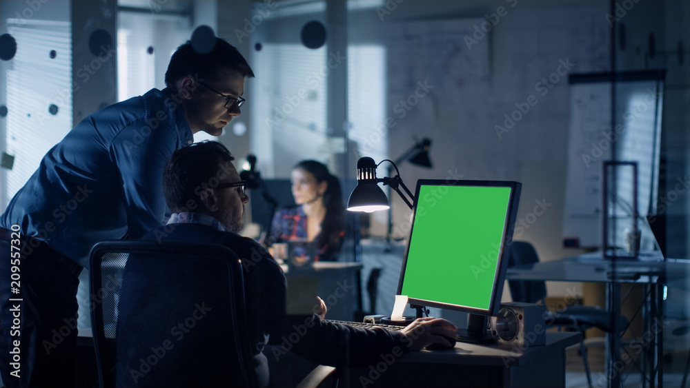 Late at Night Two Men Discuss about Green Screen Template that are Shown on Display of a Personal Computer. Office Looks Modern with Lots of Blueprints on the Walls. In the Background Woman Working. Late at Night Two Men Discuss about Green Screen Template that are Shown on Display of a Personal Computer. Office Looks Modern with Lots of Blueprints on the Walls. In the Background Woman Working.