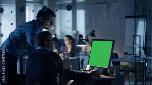 Late at Night Two Men Discuss about Green Screen Template that are Shown on Display of a Personal Computer. Office Looks Modern with Lots of Blueprints on the Walls. In the Background Woman Working.