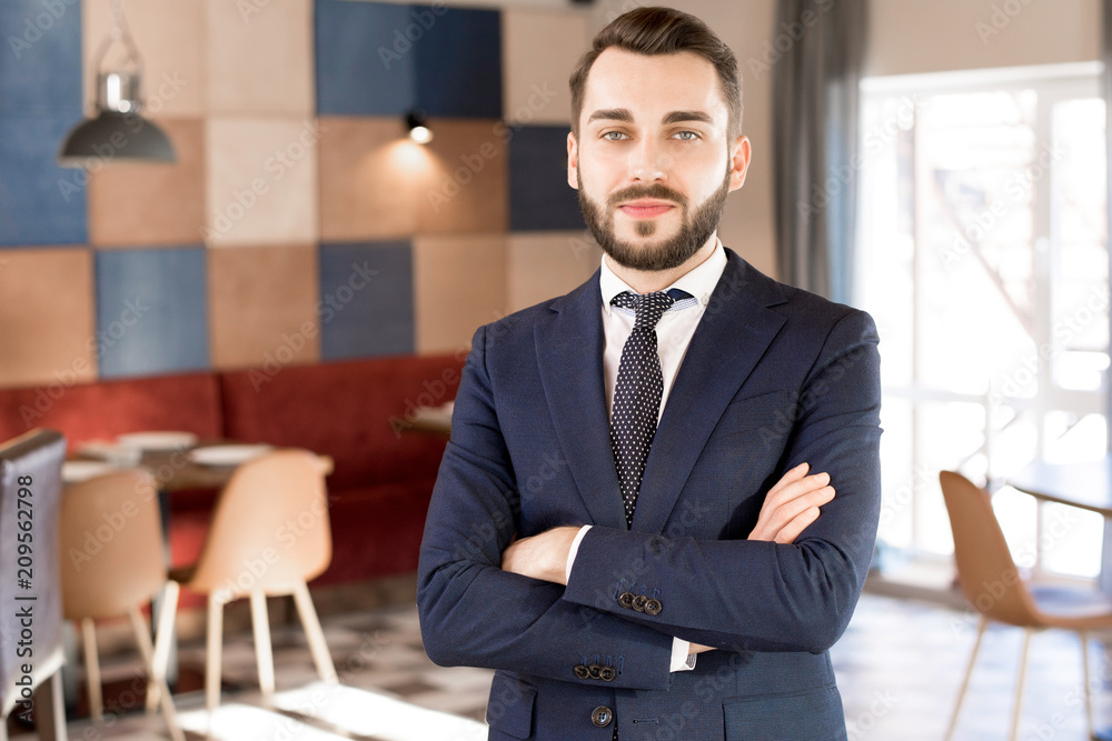 Content positive handsome young businessman with beard and mustache wearing formal jacket and tie crossing arms on chest and looking at camera in restaurant Content positive handsome young businessman with beard and mustache wearing formal jacket and tie crossing arms on chest and looking at camera in restaurant
