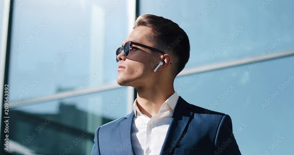 Attractive young businessman using wireless earphones standing outside. He wearing sunglasses and looks away. Communication, finance, business people, interns, CEO. Close up. Shot on Red Epic Attractive young businessman using wireless earphones standing outside. He wearing sunglasses and looks away. Communication, finance, business people, interns, CEO. Close up. Shot on Red Epic