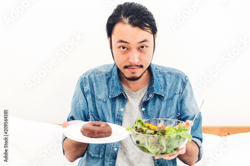 Man holding and making choice between healthy salad and calorie bomb chocolate donut on bed at home.Healthy eating and Junk food concept