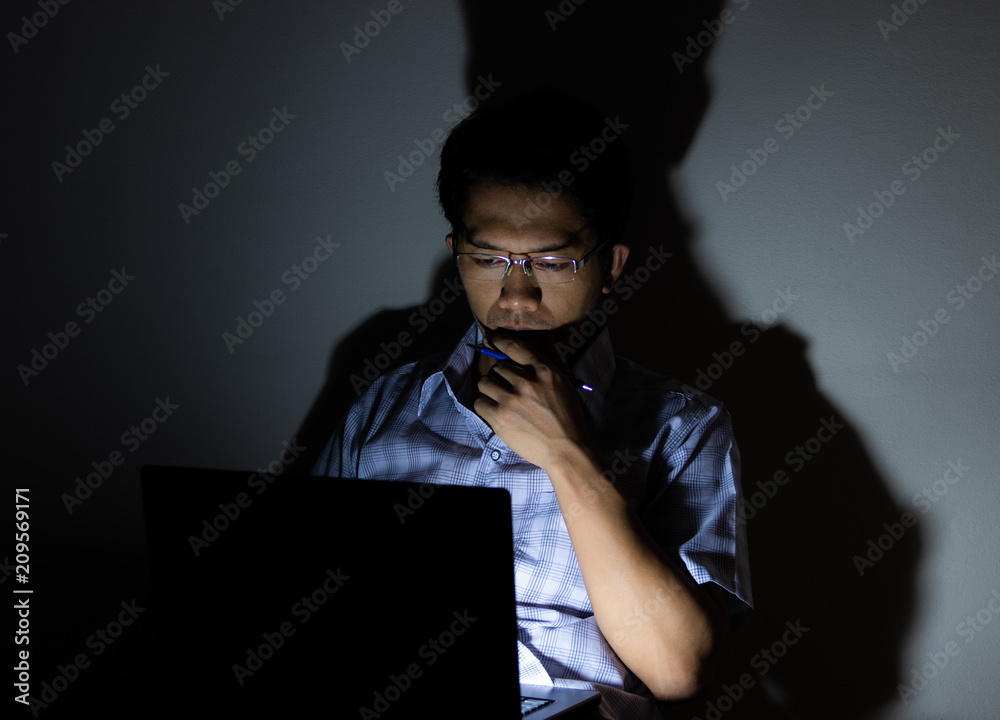 Low light view and shadows. Asian men are using a laptop and emotional are seriously. And a pen in hand. Low light view and shadows. Asian men are using a laptop and emotional are seriously. And a pen in hand.