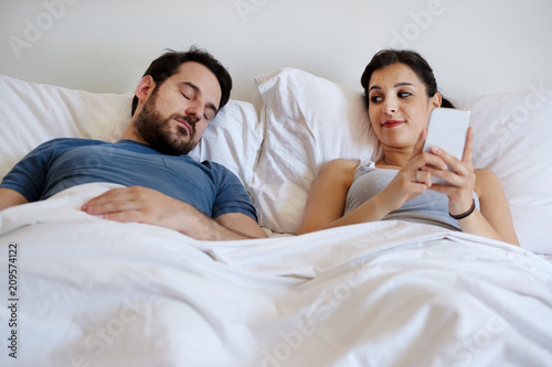 Fototapete Cheating wife using mobile phone lying in bed next to his sleeping husband