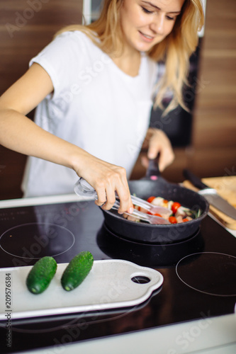 Closeup of blonde caucasian woman roasting meat kebabs on frying pan in kitchen interior.