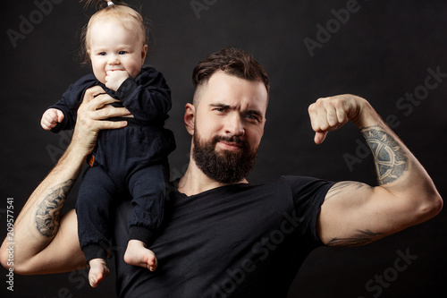 Muscular father with tattooed arms holding his little baby daughter on one shoulder and toughening mucles, demonstrating size of his biceps over black background. photo