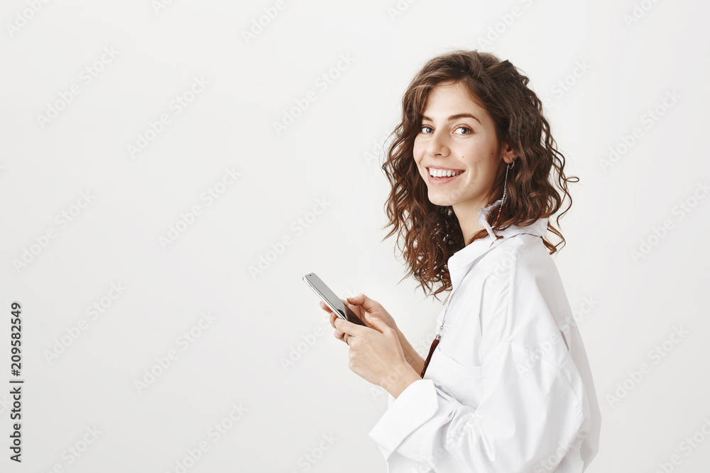 Portrait of successful caucasian businesswoman standing in profile, holding smartphone and smiling at camera, expressing positive emotions over gray background. Bloger shares thoughts with audience Portrait of successful caucasian businesswoman standing in profile, holding smartphone and smiling at camera, expressing positive emotions over gray background. Bloger shares thoughts with audience