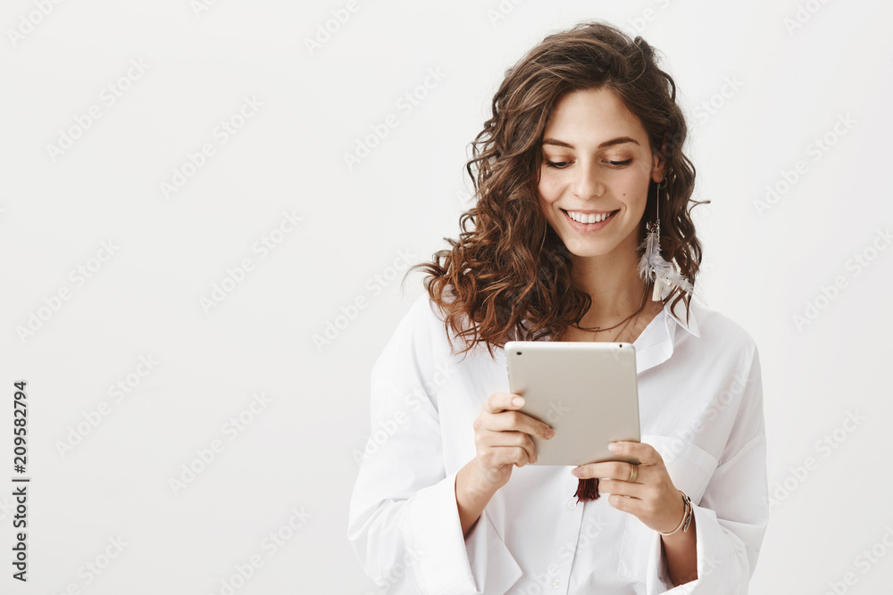 Charming cute caucasian woman in white trendy blouse browsing in net via tablet, smiling and expressing happiness, standing against gray background. Fashionable woman editing her photos in app Charming cute caucasian woman in white trendy blouse browsing in net via tablet, smiling and expressing happiness, standing against gray background. Fashionable woman editing her photos in app