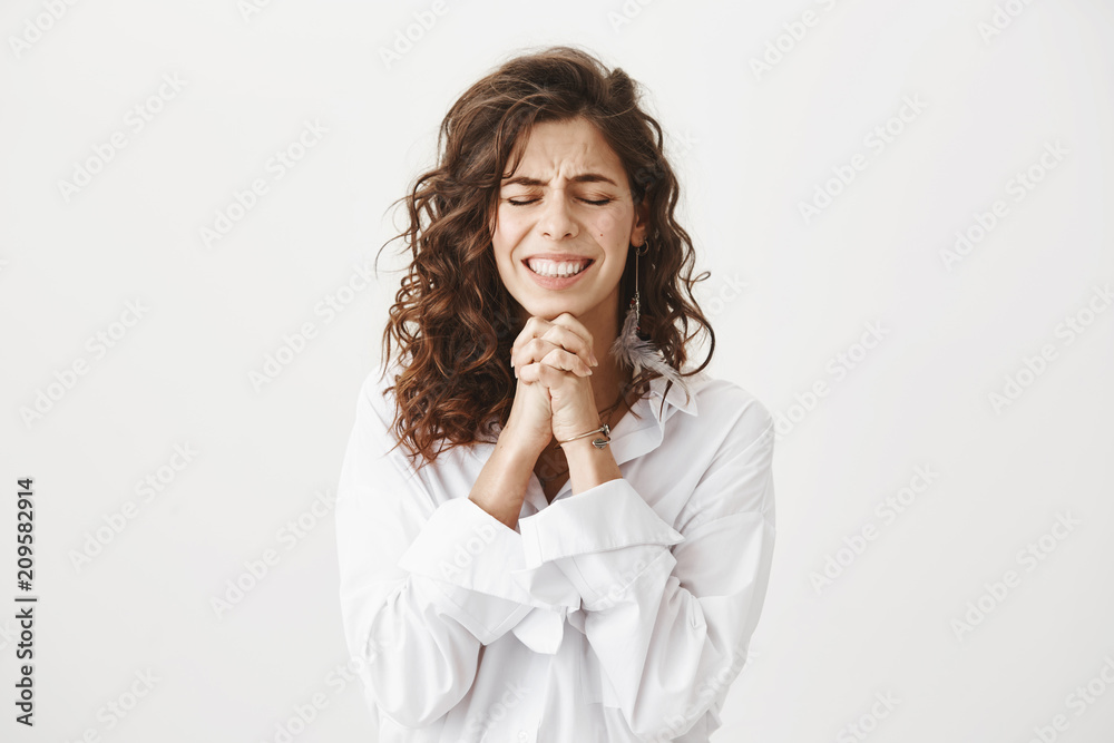 Studio shot of worried and upset caucasian woman holding hands in pray near chin, squeezing it tight and begging for something, smiling nervously and standing with closed eyes over gray background Studio shot of worried and upset caucasian woman holding hands in pray near chin, squeezing it tight and begging for something, smiling nervously and standing with closed eyes over gray background