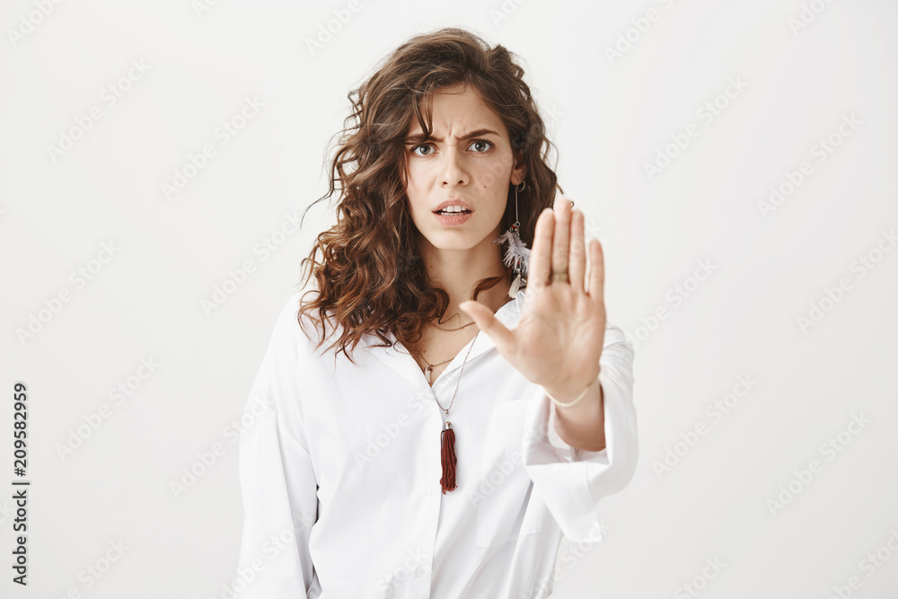 Unhappy gorgeous caucasian woman with curly brown hair showing stop or enough gesture with hand, frowning and expressing dislike and wariness, standing against gray background. Do not come closer Unhappy gorgeous caucasian woman with curly brown hair showing stop or enough gesture with hand, frowning and expressing dislike and wariness, standing against gray background. Do not come closer