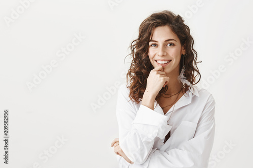 Φωτογραφία Emotive charming caucasian woman with curly hair and feather earring holding hand on ching and smiling broadly, expressing interest and positive attitude, standing against gray background