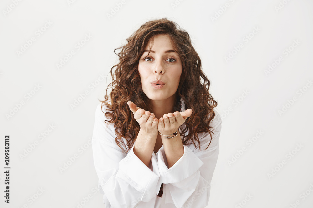 Charming caucasian woman bending towards camera with folded lips and palms near face as if blowing kiss, standing against gray background. Fashionable hairdresser sends love to her coworker Charming caucasian woman bending towards camera with folded lips and palms near face as if blowing kiss, standing against gray background. Fashionable hairdresser sends love to her coworker