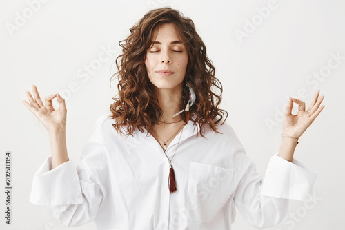 Indoor portrait of calm beautiful caucasian woman meditating in white blouse, smiling while raising hands with zen gestures, standing against gray background. Girlfriend dragged girl to yoga classes