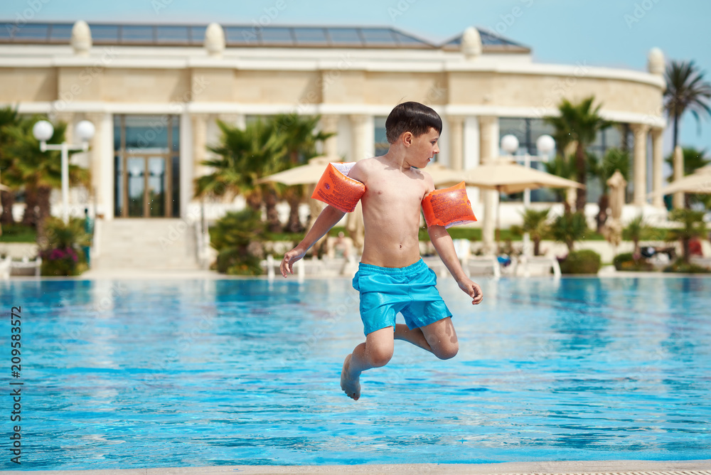 Caucasian boy in floating sleeves jumping into water in swimming pool at resort. He is spinning in air.