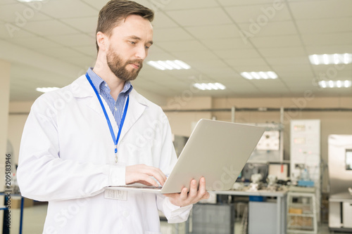 Concentrated middle-aged inspector wearing lab coat standing at spacious production department of modern factory and writing review on laptop after conducted quality control of goods. photo