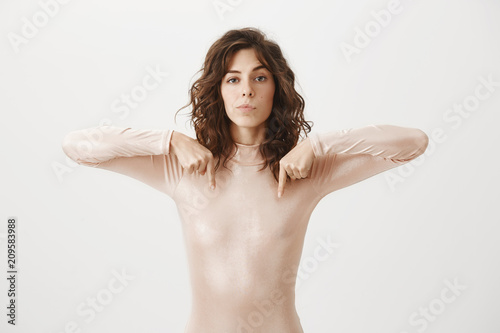 Studio shot of confident serious-looking female in beige outfit, pointing down, having no care what people think, standing self-assured over gray background as if naked, feeling ok show nipples