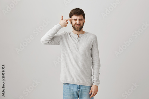 I would better kill myself than do paperwork. Portrait of tired playful european office worker showing gun sign near temple, closing eyes and squinting as if shooting, posing over gray background