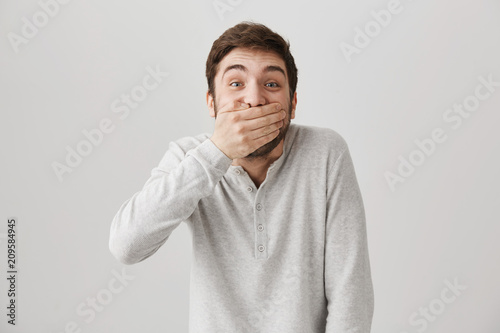 Emotive positive young male covering mouth to stop laughter or hide smile, hearing or seeing something hilarious while standing over gray background. Man can not stop lauihing over favorite tv show