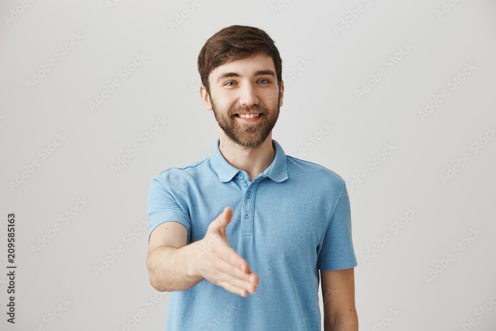 Pleased to finally meet you. Cheerful good-looking male with beard pulling palm towards camera to give handshake, standing over gray background with friendly smile while getting to know new staff Pleased to finally meet you. Cheerful good-looking male with beard pulling palm towards camera to give handshake, standing over gray background with friendly smile while getting to know new staff