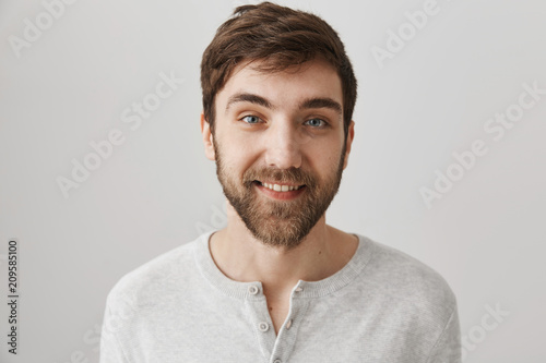 Close-up studio portrait of friendly good-looking male with beard, wearing white pullover and standing over gray background, smiling joyfully at camera. Helpful employee ready to work hard