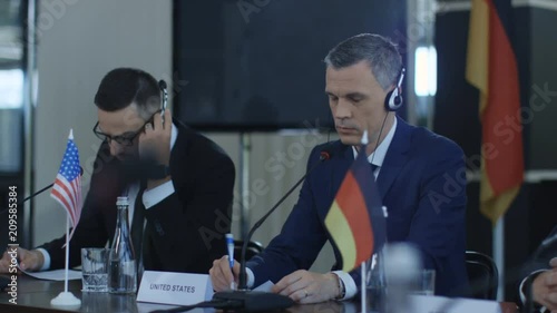 Representative men of different countries sitting at table on conference and listening to speech interpretation in headphones