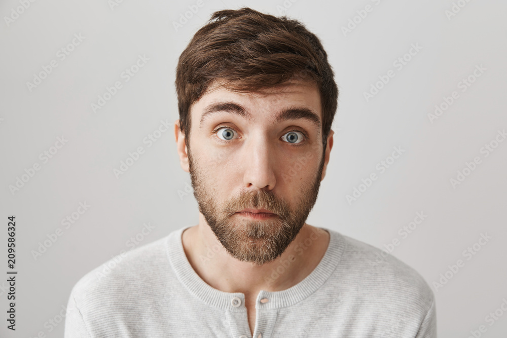 Obraz premium Close-up portrait of surprised and upset european male with beard standing with lifted eyebrows, clueless and tired expression over gray background. Guy is shocked to hear sad news