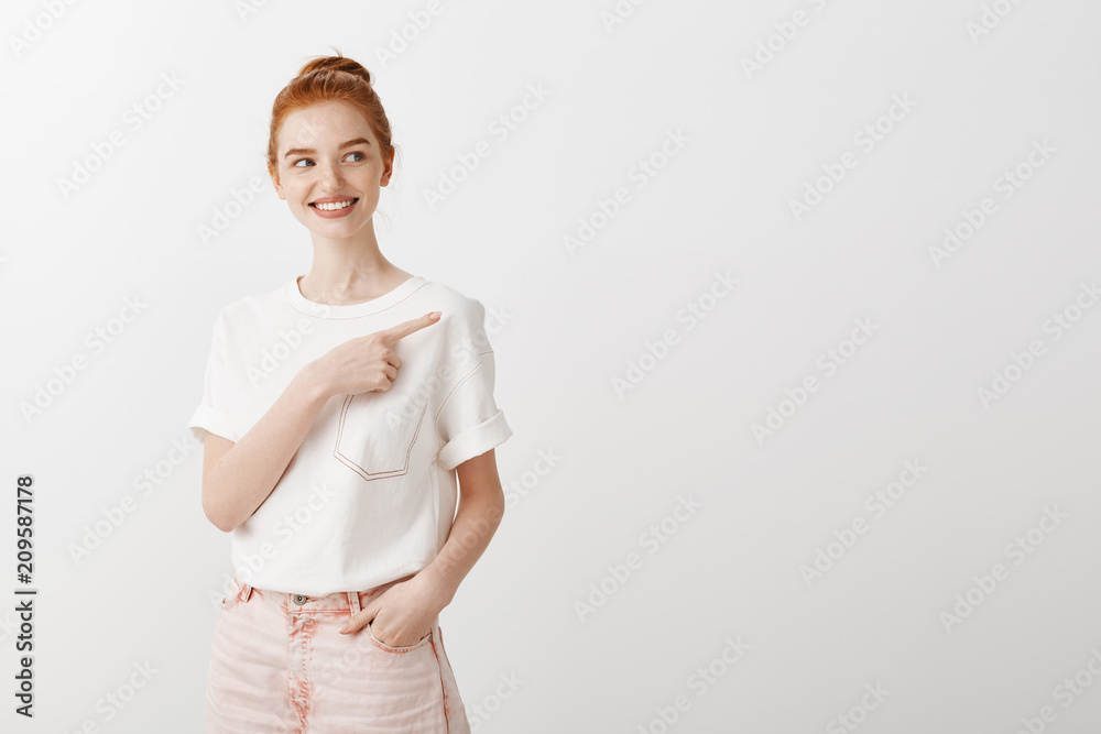 Portrait of attractive carefree female ginger girl in stylish t-shirt and jeans, holding hand in pocket and pointing right and gazing with intrigued and broad smile, posing over gray background Portrait of attractive carefree female ginger girl in stylish t-shirt and jeans, holding hand in pocket and pointing right and gazing with intrigued and broad smile, posing over gray background