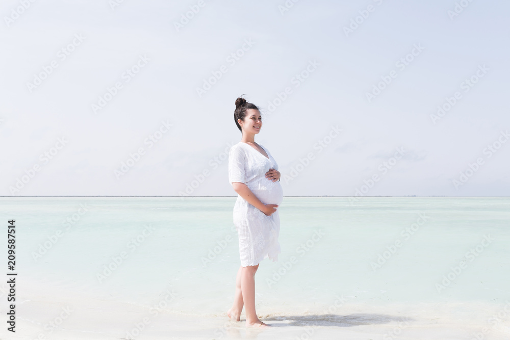 Young pregnant woman in white dress standing on the beach near blue sea and breething. Summer vacation during pregnancy, happy motherhood concept Young pregnant woman in white dress standing on the beach near blue sea and breething. Summer vacation during pregnancy, happy motherhood concept