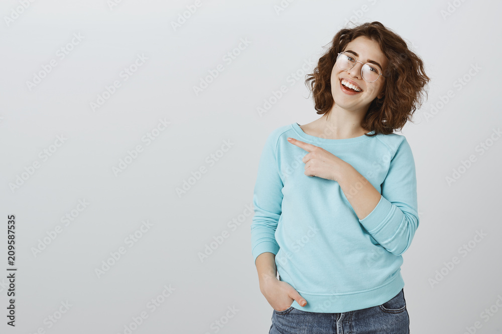 Suggesting friend to grab there cup of coffee. Portrait of satisfied happy caucasian female student in glasses, tilting head with carefree attitude, pointing at upper left corner and smiling broadly Suggesting friend to grab there cup of coffee. Portrait of satisfied happy caucasian female student in glasses, tilting head with carefree attitude, pointing at upper left corner and smiling broadly