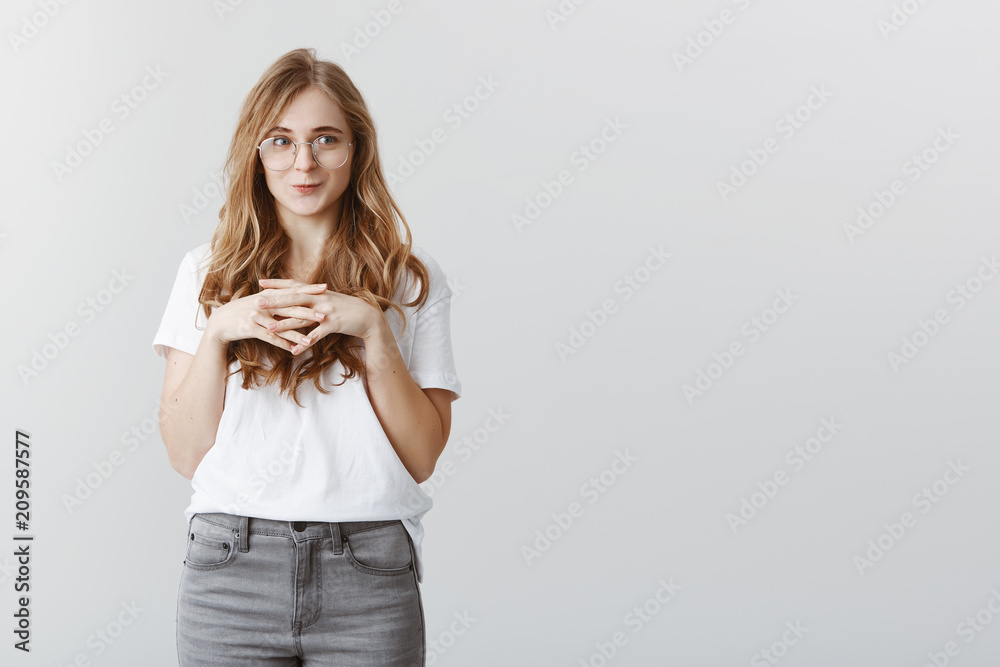 Cute coworker hesitating to confess to colleague. Timid girl in transparent glasses and casual clothes, holding hands together and making unconfident tender face, smiling silly, having no courage