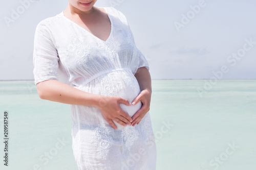 Young pregnant woman in white dress standing on the beach near blue sea and breething. Summer vacation during pregnancy, happy motherhood concept