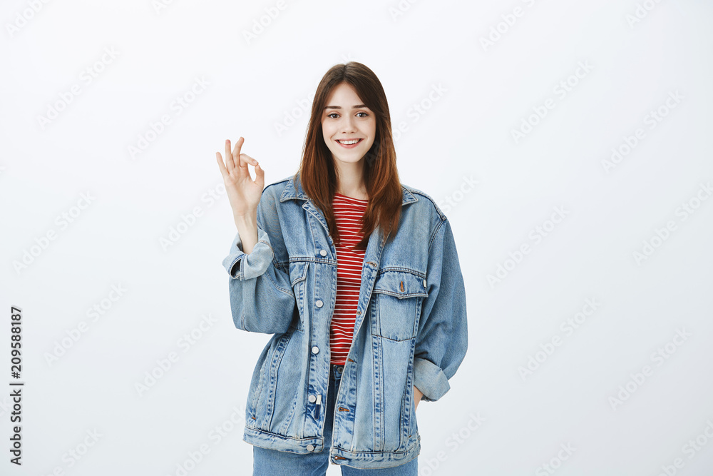 Creative smart girl showing she can deal with anything. Portrait of happy carefree confident woman in stylish denim jacket, holding hand in pocket and showing okay or excellent gesture over gray wall