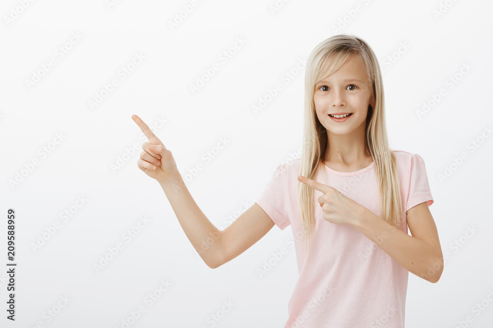 Active adorable female child with blond hair in trendy pink t-shirt, pointing at upper left corner and smiling broadly, asking parents permission to ride on ferris wheel, standing over gray background Active adorable female child with blond hair in trendy pink t-shirt, pointing at upper left corner and smiling broadly, asking parents permission to ride on ferris wheel, standing over gray background