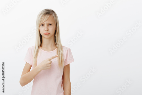 Girl upset and jealous  wanting new toy. Portrait of sad displeased gloomy child with blond hair in pink t-shirt  pointing at upper right corner  pouting and feeling offended  whining over gray wall