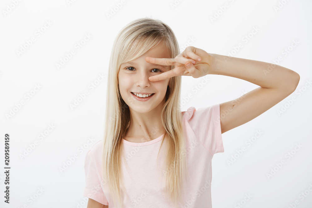 Waist-up shot of positive charming child with blond hair in casual outfit, showing victory or peace gesture over eye and smiling happily, dancing or having fun over gray wall, being in great mood