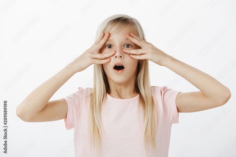 Indoor portrait of shocked surprised adorable blond girl in pink t-shirt, dropping jaw, saying wow while holding fingers near eyes, staring at camera after knowing amazing news, gasping over gray wall Indoor portrait of shocked surprised adorable blond girl in pink t-shirt, dropping jaw, saying wow while holding fingers near eyes, staring at camera after knowing amazing news, gasping over gray wall