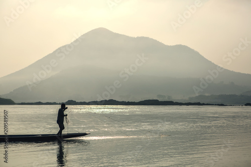  Thai people is highly skilled fishermen. Man stands on the end of long narrow pirogues to cast his nets. View of mountain and river, natural background.