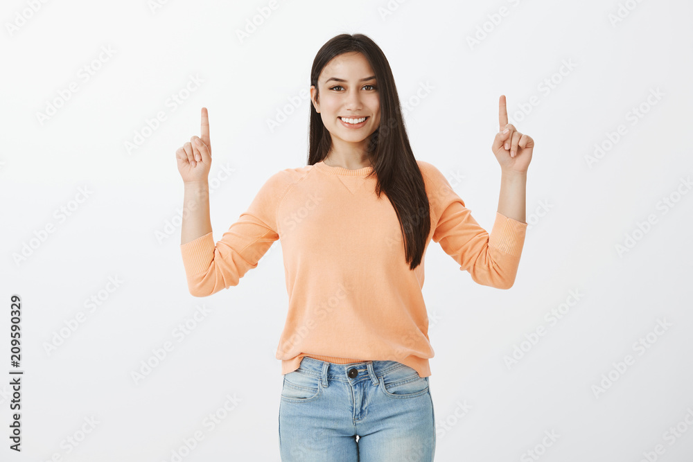 Satisfied happy female student in pullover and jeans raising index fingers and pointing up with index fingers while smiling cheerfully and gazing at camera, showing great copy space Satisfied happy female student in pullover and jeans raising index fingers and pointing up with index fingers while smiling cheerfully and gazing at camera, showing great copy space