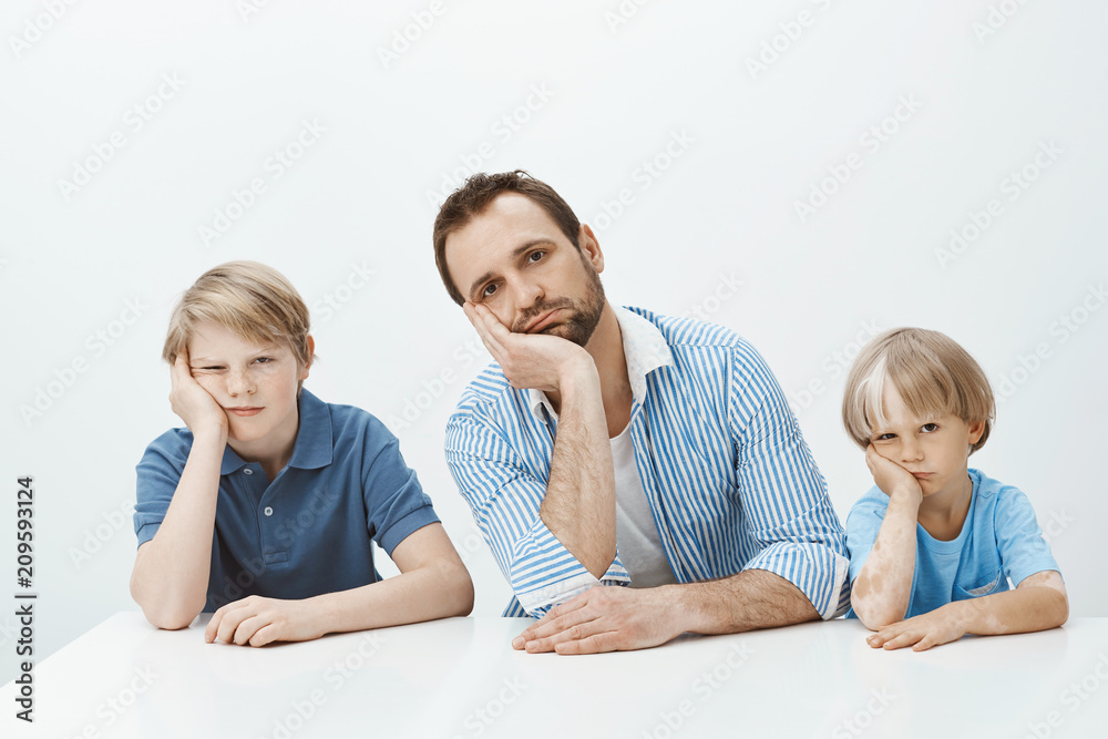 Boys feeling bored and upset. Portrait of tired funny european family of sons and dad sitting at table, leaning heads on palms and staring indifferent at camera, being careless while mom scolding them Boys feeling bored and upset. Portrait of tired funny european family of sons and dad sitting at table, leaning heads on palms and staring indifferent at camera, being careless while mom scolding them