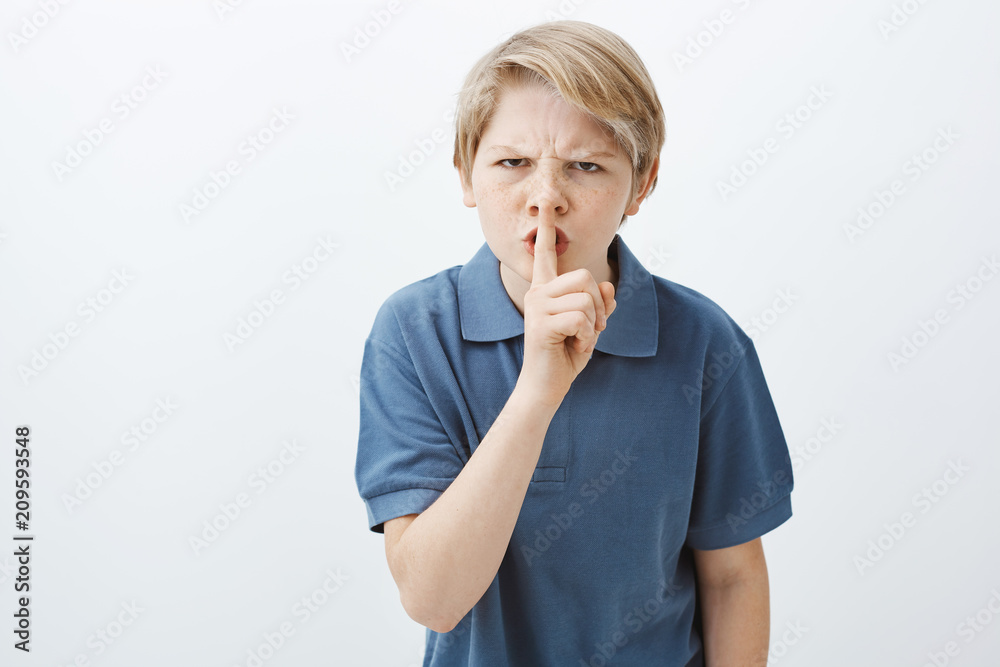 Indoor shot of angry annoyed blond brother in casual t-shirt, frowning,  saying shh while making shush gesture with index finger over mouth,  demanding to stay quiet, wanting silence and peace Photos |