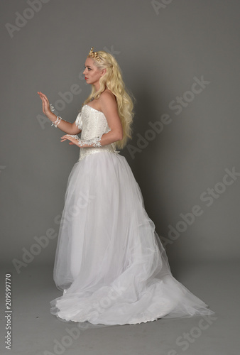 full length portrait of blonde girl wearing white fantasy gown. standing pose in side profile, grey studio background.