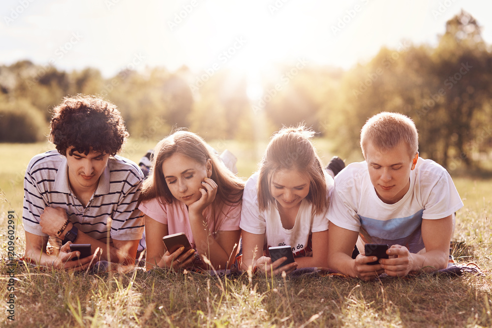 Fototapeta premium Four friends focused in their smart phones, enjoy leisure time on nature, lie on green grass, share photos after picnic in their social networks, pose against sunshine. Teenagers, technology