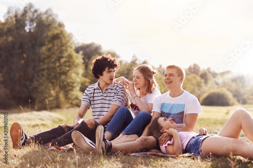Outdoor shot of happy youngsters have charming smiles, look into distance, sit on plaid, pose against green nature and sunshine background. Four friends have good realtionship, pincic outside