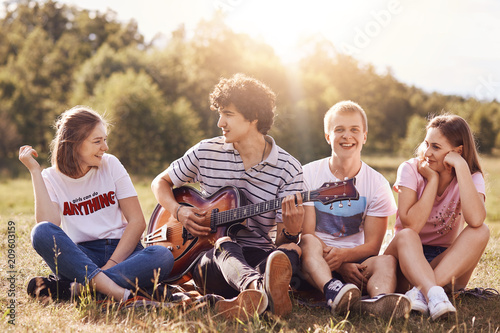 Happy young friends sing songs to guitar, have joy together, recreat outdoor, sit on green grass. Curly handsome male teenager plays guitar, enteratins his companions, enjoys summer warm day. © sementsova321
