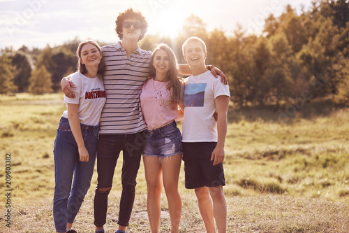 Photo of close friends stand outdoor, have picnic together, embrace and smile happily at camera, have good relationship, dressed in casual summer clothes. People, companionship, rest concept