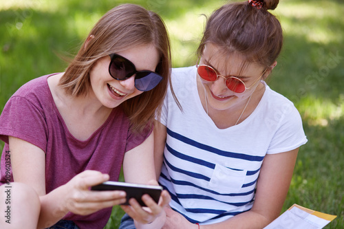 Two attractive women in trendy sunglasses have happy expressions, dressed in casual t shirts, watch funny video on smart phone, use wireless internet, pose in park against green grass background. photo