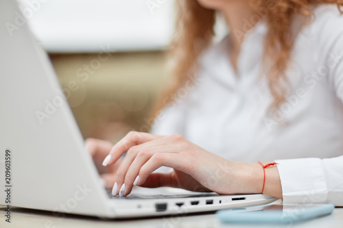 Cropped view of successful female entrepreneur sits in front of opened laptop computer, works on financial report, keyboards information, wears elegant white blouse. People, work, lifestyle concept
